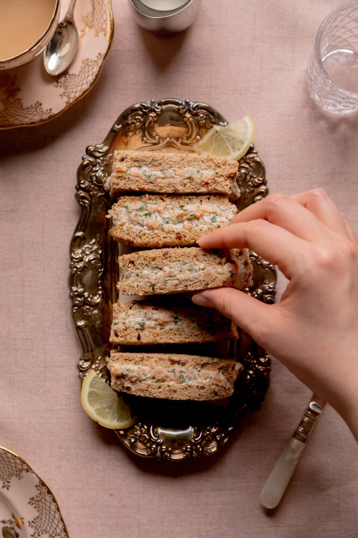 A hand grabbing a smoked salmon tea sandwich from a plate of sandwiches.