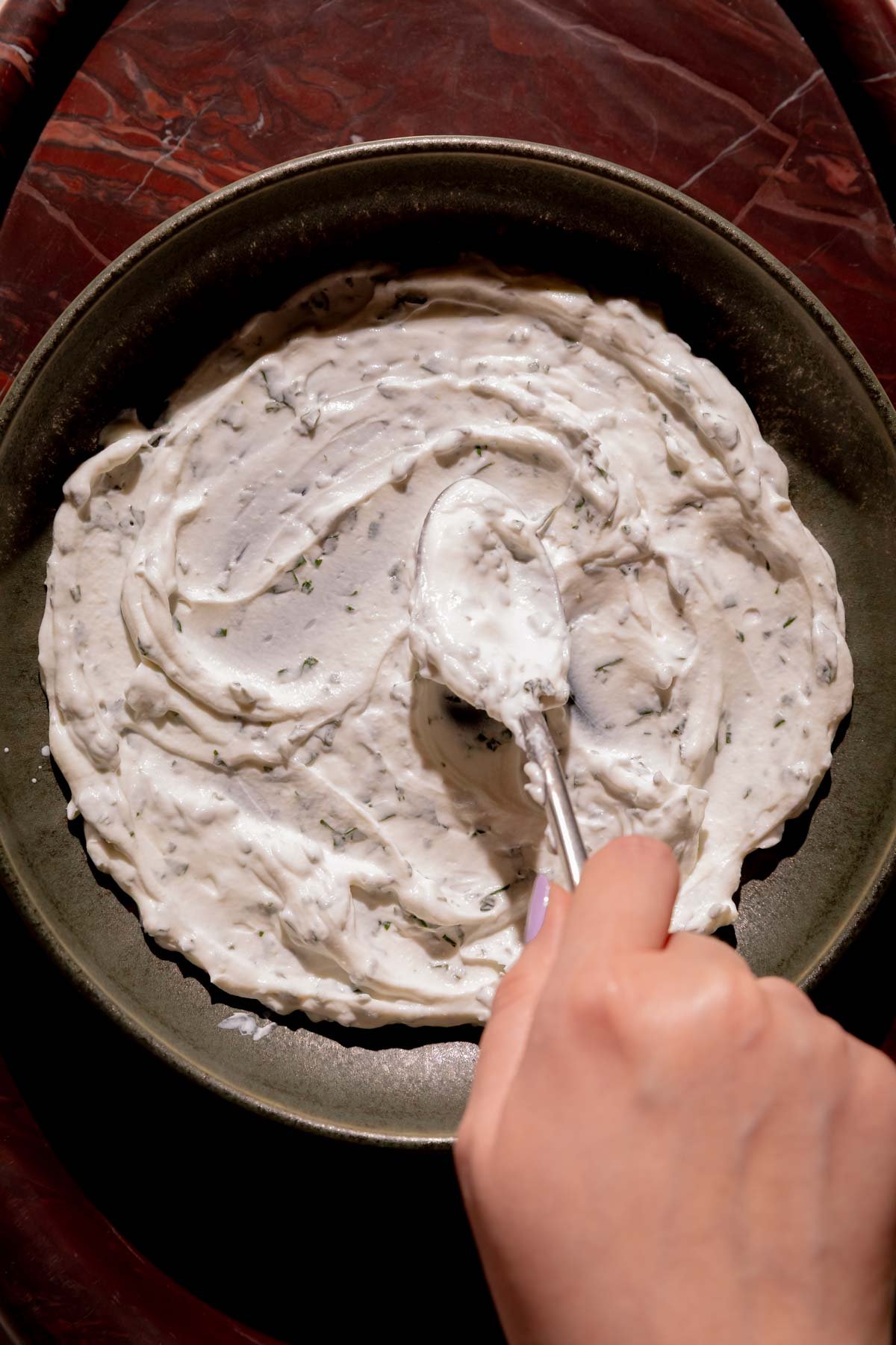 A hand swirling thai basil yogurt onto a serving plate with a spoon.