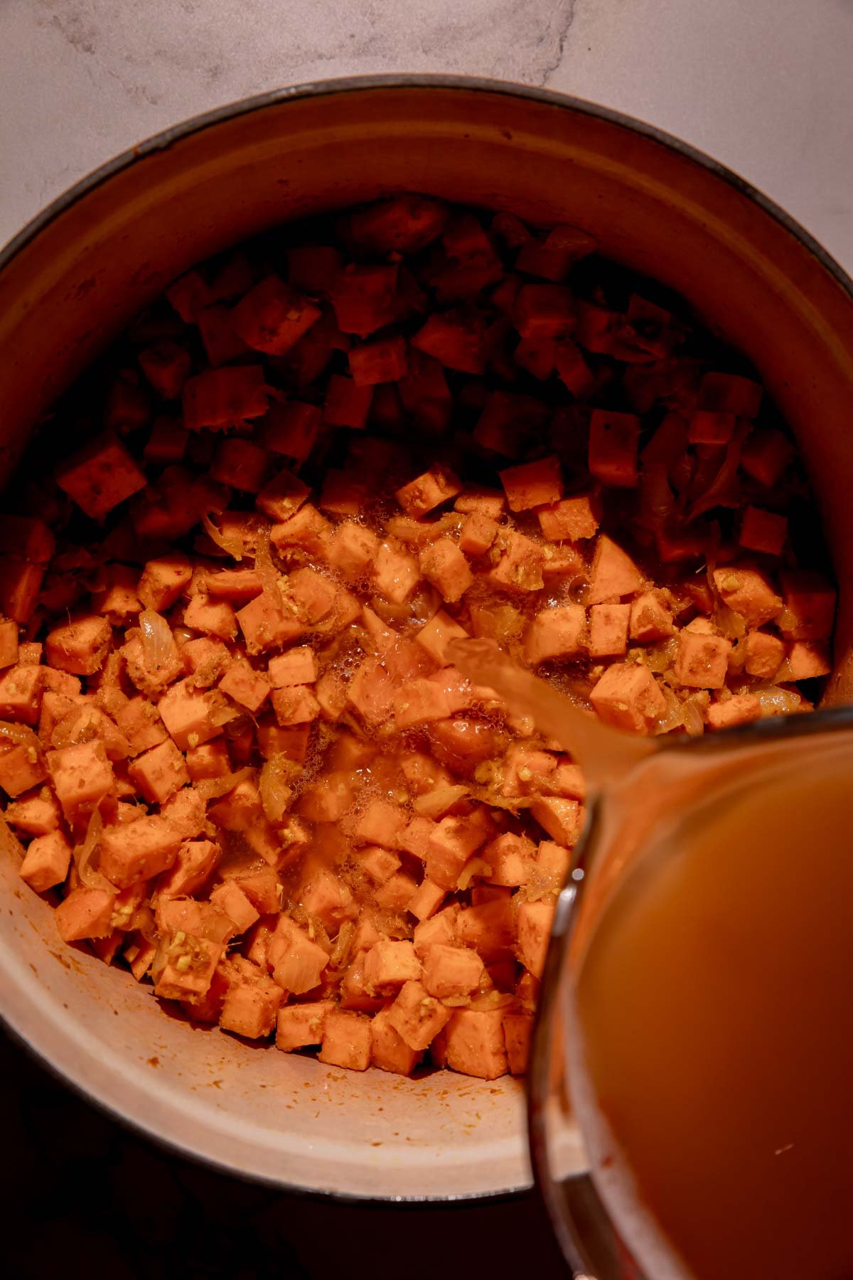 Chicken stock being poured into a Dutch oven filled with sweet potatoes.