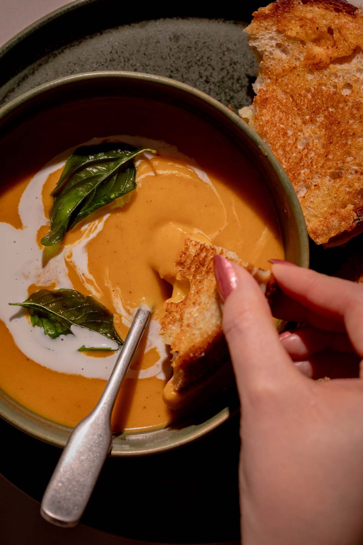A hand dipping a piece of grilled cheese into a bowl of curried coconut sweet potato soup.