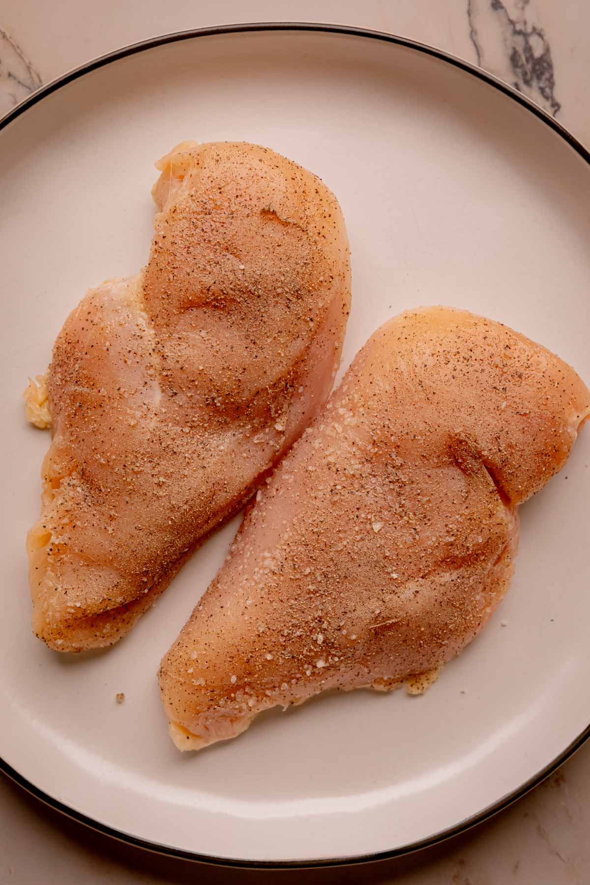 Raw chicken breasts sprinkled with kosher salt and ground black pepper.