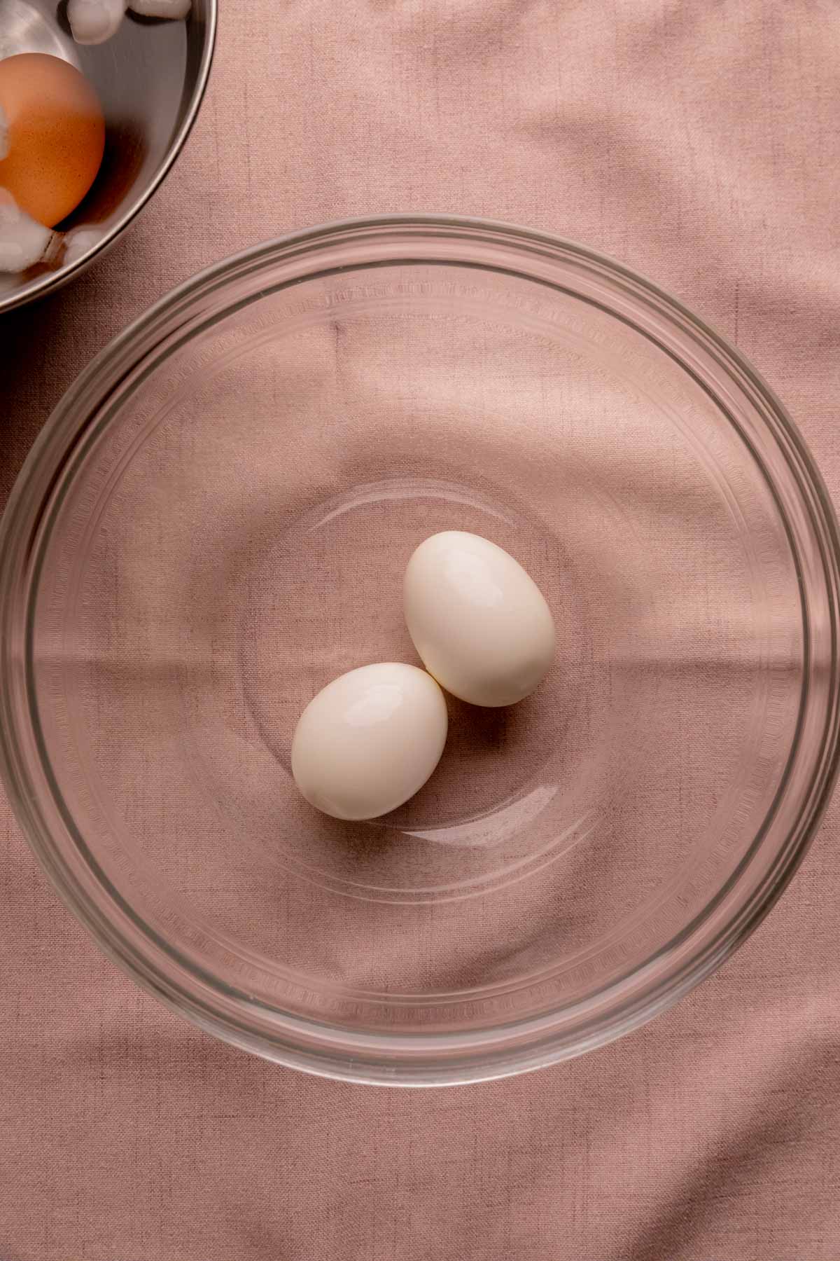 A bowl of peeled, boiled eggs next to a bowl of ice water with an unpeeled egg in it.